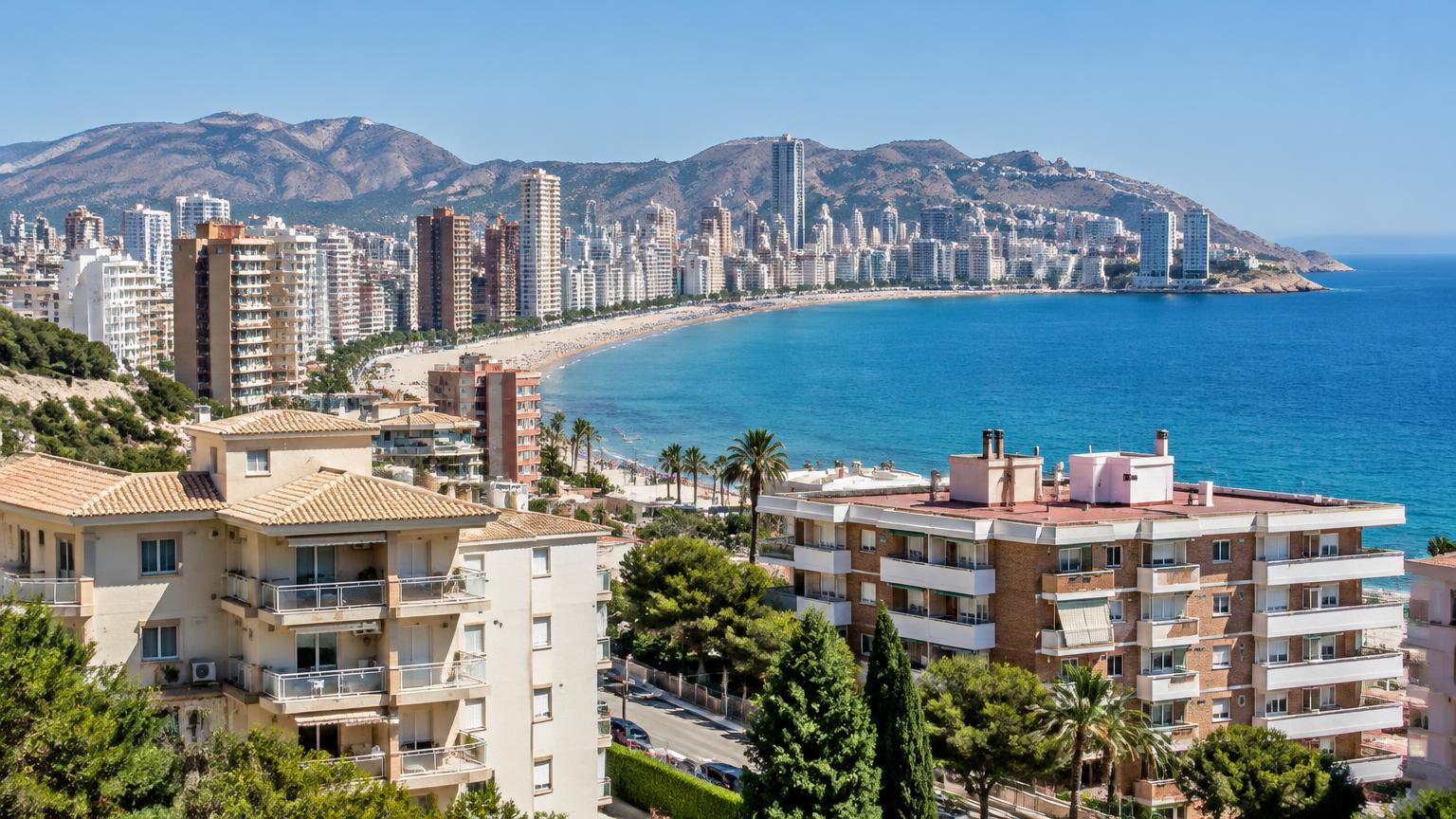 Blick über Wohnanlagen auf die Bucht von Benidorm mit Hochhäusern, Strand und Bergen.